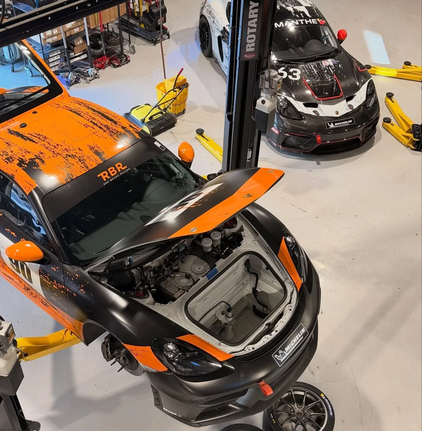 Red Beard Racing's orange and black Porsche 718 Cayman GT4 with its hood open in the workshop